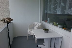 Ein kleiner Balkon mit einem weißen Plastikstuhl und einem weißen Tisch, eine Topfpflanze auf dem Tisch und ein Fenster mit einem blumigen Vorhang.