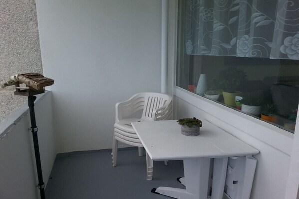 A small balcony with a white plastic chair and a white table, a potted plant on the table, and a window with a floral curtain. - Holiday apartment with 2 bedrooms (Goslar\/Hahnenklee)