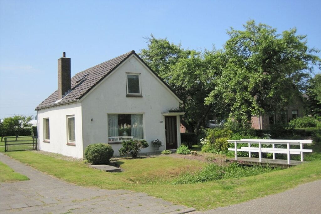 A single-story house with a white exterior, a chimney, and a small front yard with a bench.