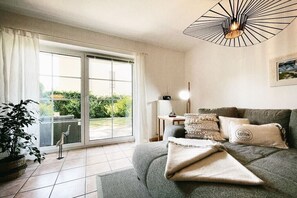 Ein Wohnzimmer mit Sofa, einer Lampe, einer Pflanze und Blick durch eine Glastür.