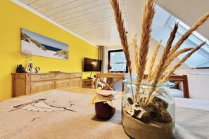 A room with a yellow wall, a painting, a television, a table with a glass jar and a small pot, and a window with a view. - Holiday apartment with 1 bedroom (Dierhagen)