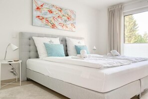 A bedroom with a bed, a nightstand, a lamp, a window with curtains, and a wall painting. - House on the beach, Apartment 1 (Scharbeutz)