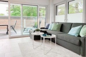 A modern living room with a grey sofa, white coffee table, and a glass door leading to an outdoor area. - House on the beach, Apartment 1 (Scharbeutz)