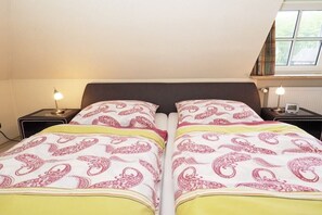 A double bed with patterned bedding, two bedside tables with lamps, and a window with a view of greenery. - Holiday apartment with 2 bedrooms (Wyk auf Föhr)