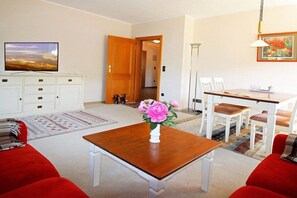 A living room with a television, a coffee table with a vase of flowers, a dining area with chairs, and a hallway leading to another room. - Holiday apartment with 2 bedrooms (Wyk auf Föhr)