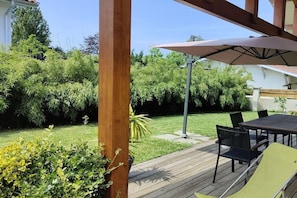 Una terraza de madera con mesa y sillas, rodeada de abundante vegetación y un cielo despejado.