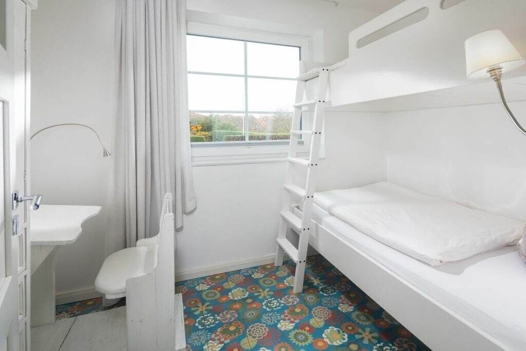 A small, white room with a bunk bed, a window with curtains, a sink, and a colorful patterned floor.