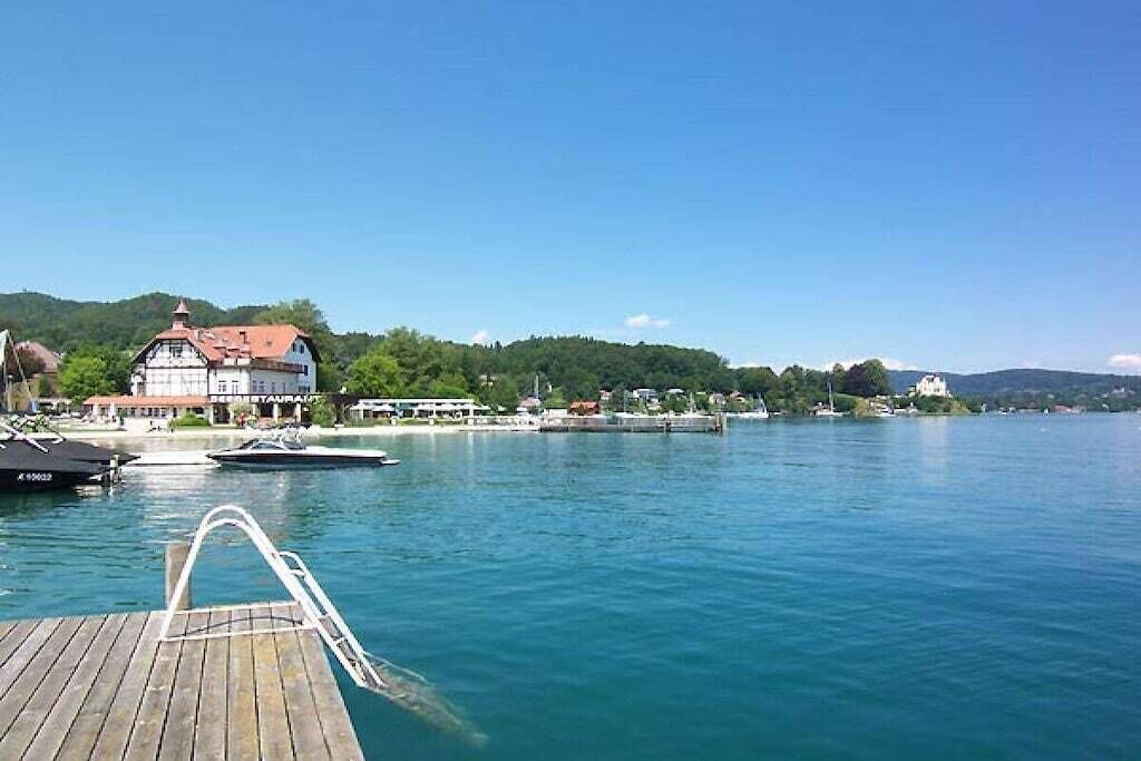 Un muelle de madera que conduce a una masa de agua con barcos, un edificio con techo rojo y un paisaje montañoso al fondo.