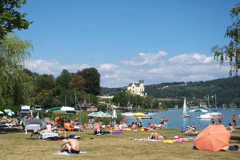 Una playa junto al lago con gente tomando el sol, barcos y un castillo al fondo.