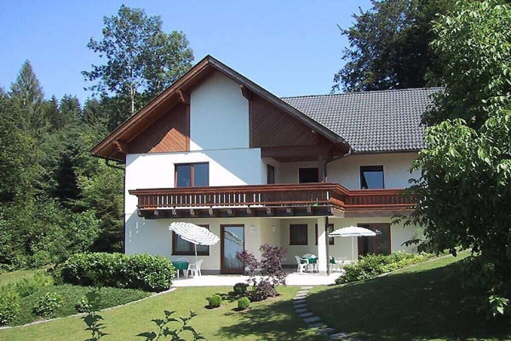 Una casa de dos pisos con un balcón, un patio cubierto con mobiliario exterior y un jardín cuidado.
