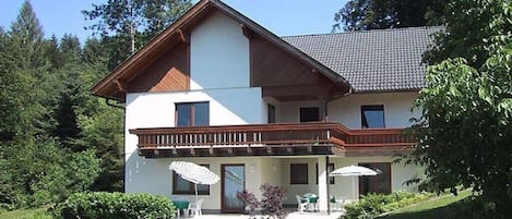 Una casa de dos pisos con un balcón, un patio cubierto con mobiliario exterior y un jardín cuidado.