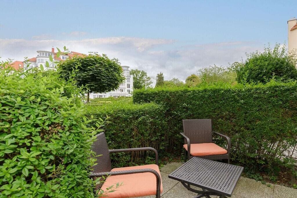 An outdoor seating area with wicker chairs and a metal table, surrounded by greenery and a hedge.