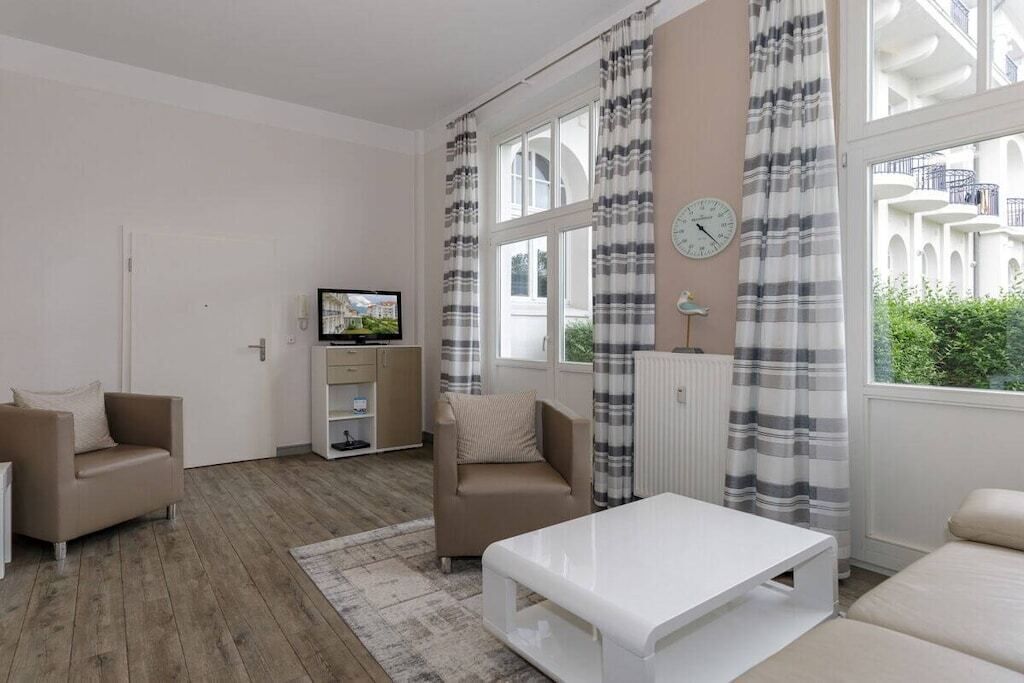 Ein modernes Wohnzimmer mit einer Couch, einem Couchtisch und einem Fernseher. An den Fenstern hängen Vorhänge, und an der Wand ist eine Uhr zu sehen.
