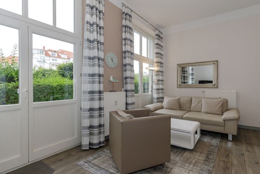 A room with a sofa, a chair, a small table, a clock, a mirror, and large windows with curtains.