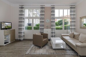 A living room with a sofa, armchair, coffee table, TV, and curtains. - Holiday apartment Haus am Park 322251 (Kühlungsborn)