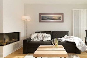 A modern living room with a black sofa, a wooden coffee table, a fireplace, and a framed picture on the wall. - Holiday apartment Villa Löwenstein 319571 (Kühlungsborn)