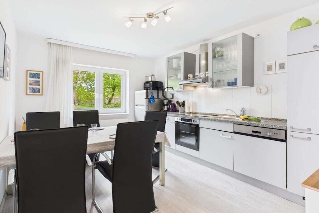 Eine moderne Küche mit weißen Schränken, ein Essbereich mit Tisch und Stühlen sowie ein Fenster mit Blick auf Grünflächen.