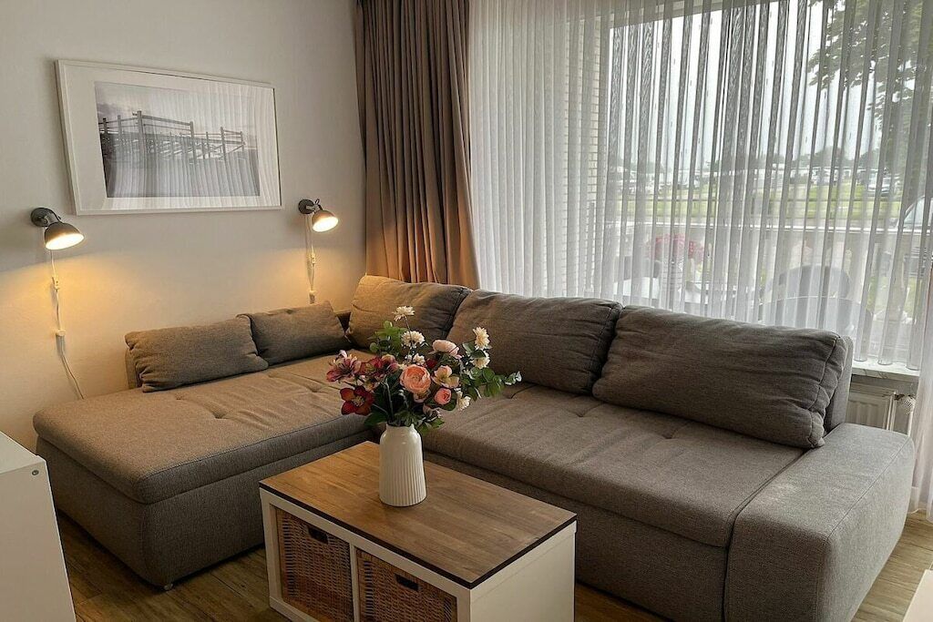 A living room with a sectional sofa, a small wooden coffee table with a vase of flowers, two wall-mounted lamps, and a window with sheer curtains.