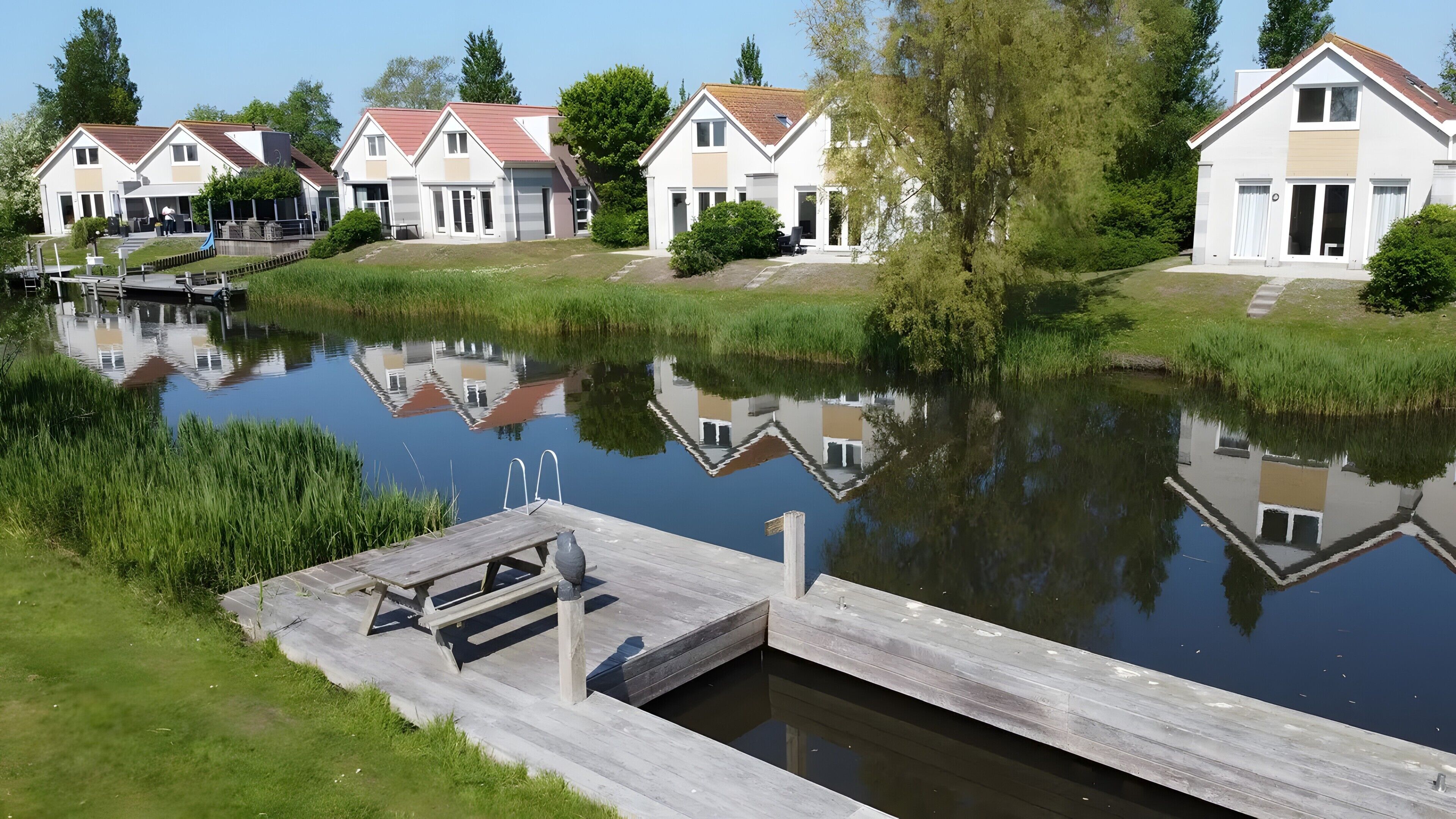 A row of modern houses with a calm water body in front, wooden dock, and greenery.