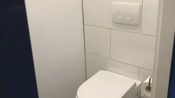 Inodoro blanco con palanca de descarga y pared blanca con interruptor de luz.