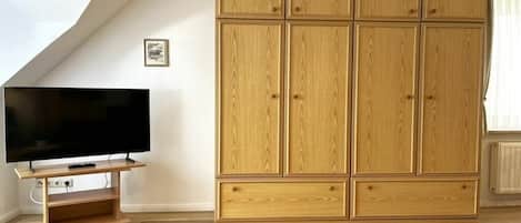 A room with a large wooden wardrobe, a flat-screen TV on a wooden stand, and a small plant on a wooden table.