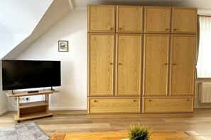 Ein Zimmer mit großem Holzschrank, einem Flachbildfernseher auf einem Holzgestell und einer kleinen Pflanze auf einem Holztisch.