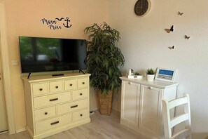 A room with a television on a cabinet, a potted plant, a clock on the wall, and a small table with a plant and a picture frame. - Holiday apartment in Büsum with swimming pool (Büsum)
