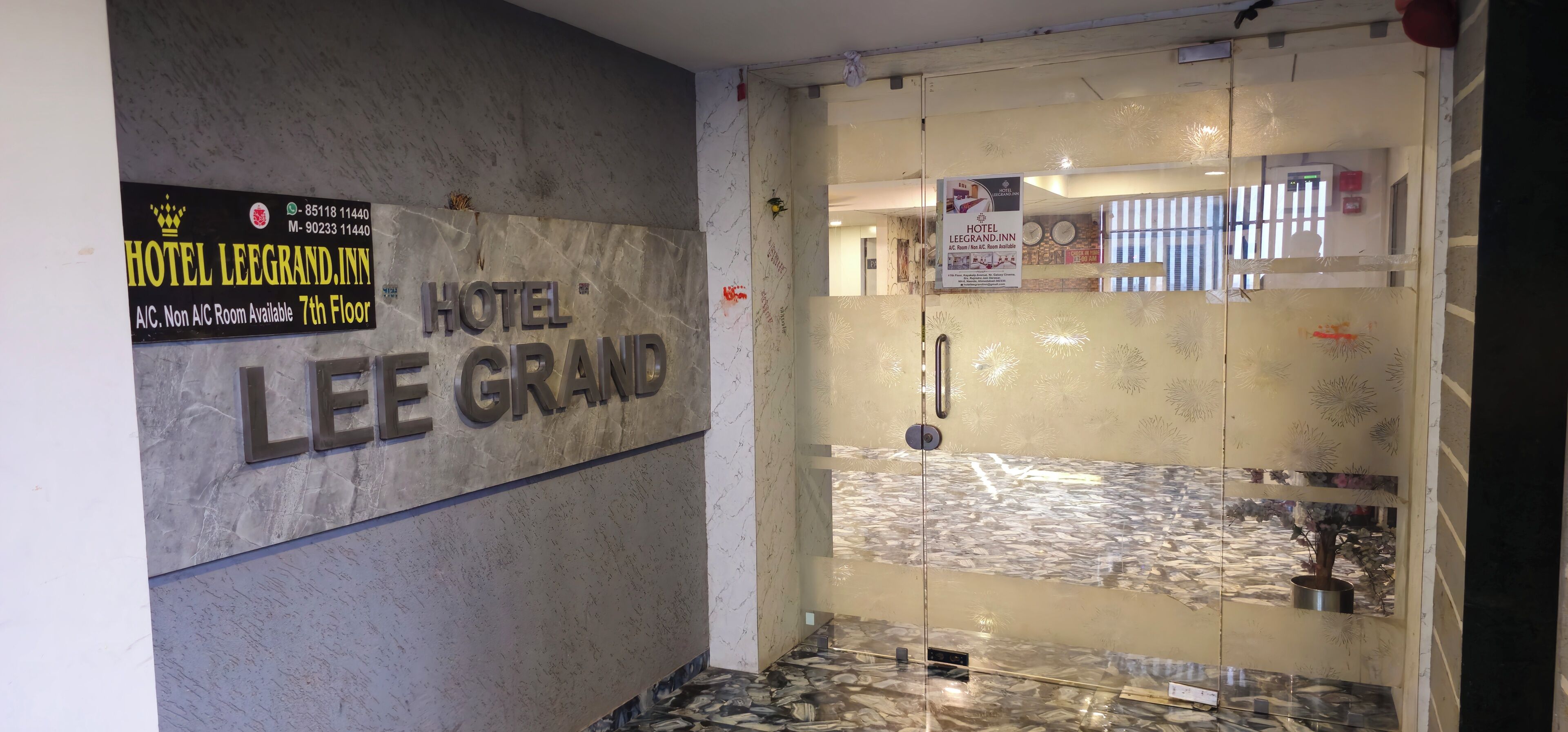Entrada de un hotel con un letrero que dice HOTEL LEE GRAND, un número de contacto y una nota sobre disponibilidad de habitaciones con aire acondicionado en el séptimo piso.