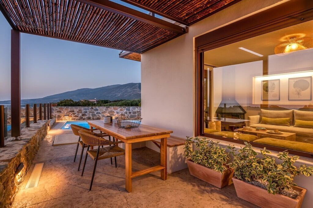 Eine Terrasse im Freien mit einem Holztisch und -stühlen, einer Steinmauer und Blick auf das Meer und die Berge.