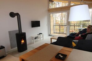 A modern living room with a wood-burning stove, a white TV, and a large window overlooking a deck. - Holiday apartment with 2 bedrooms (Crinitzberg)