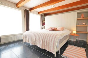 Een slaapkamer met een bed, nachtkastje, lamp, raam met gordijnen en een plank met decoratieve spullen.