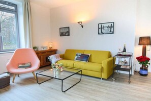 A living room with a yellow sofa, a pink chair, a coffee table, and a small side table with a lamp and a vase of flowers. - Holiday apartment with 1 bedroom (Burg auf Fehmarn)