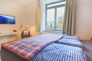 A bedroom with a bed, a chair, a TV, and a window with curtains. - Holiday apartment with 1 bedroom (Burg auf Fehmarn)