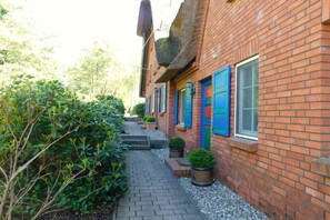 Un étroit sentier pavé de briques, bordé d’une végétation luxuriante et d’une rangée de bâtiments aux volets bleus.