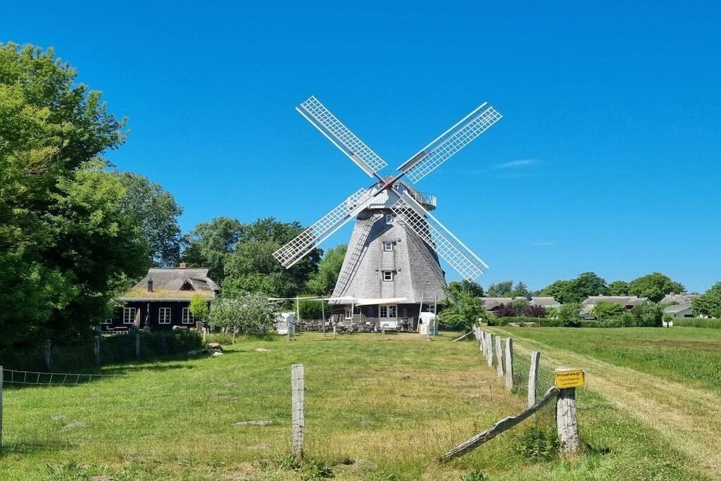 Eine traditionelle Windmühle mit einem einstöckigen Gebäude, umgeben von einem eingezäunten Feld und einem Haus im Hintergrund.