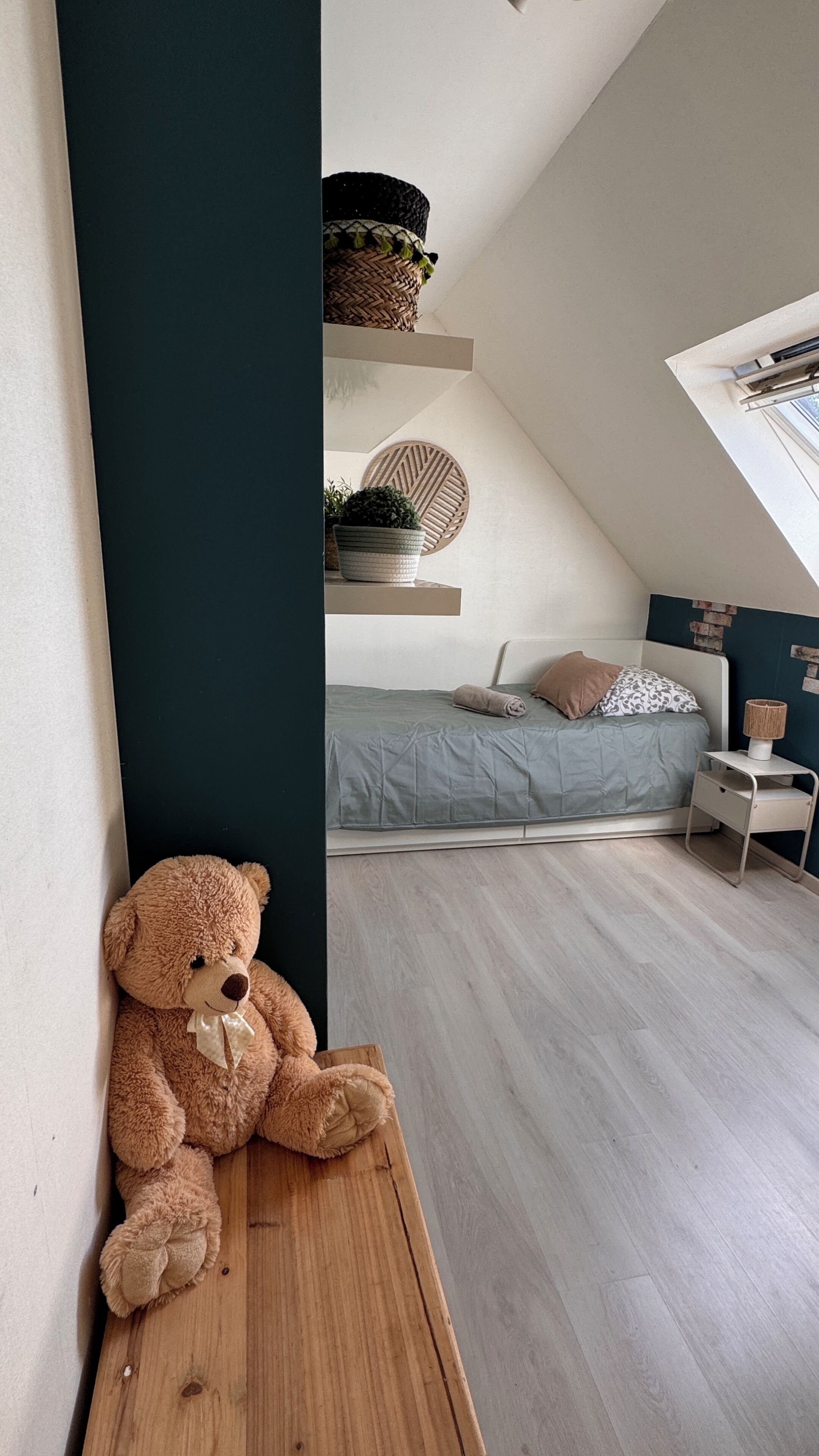 A cozy bedroom with a sloped ceiling, a wooden bench, a teddy bear, a bed with a grey bedspread, and a bedside table with a lamp.