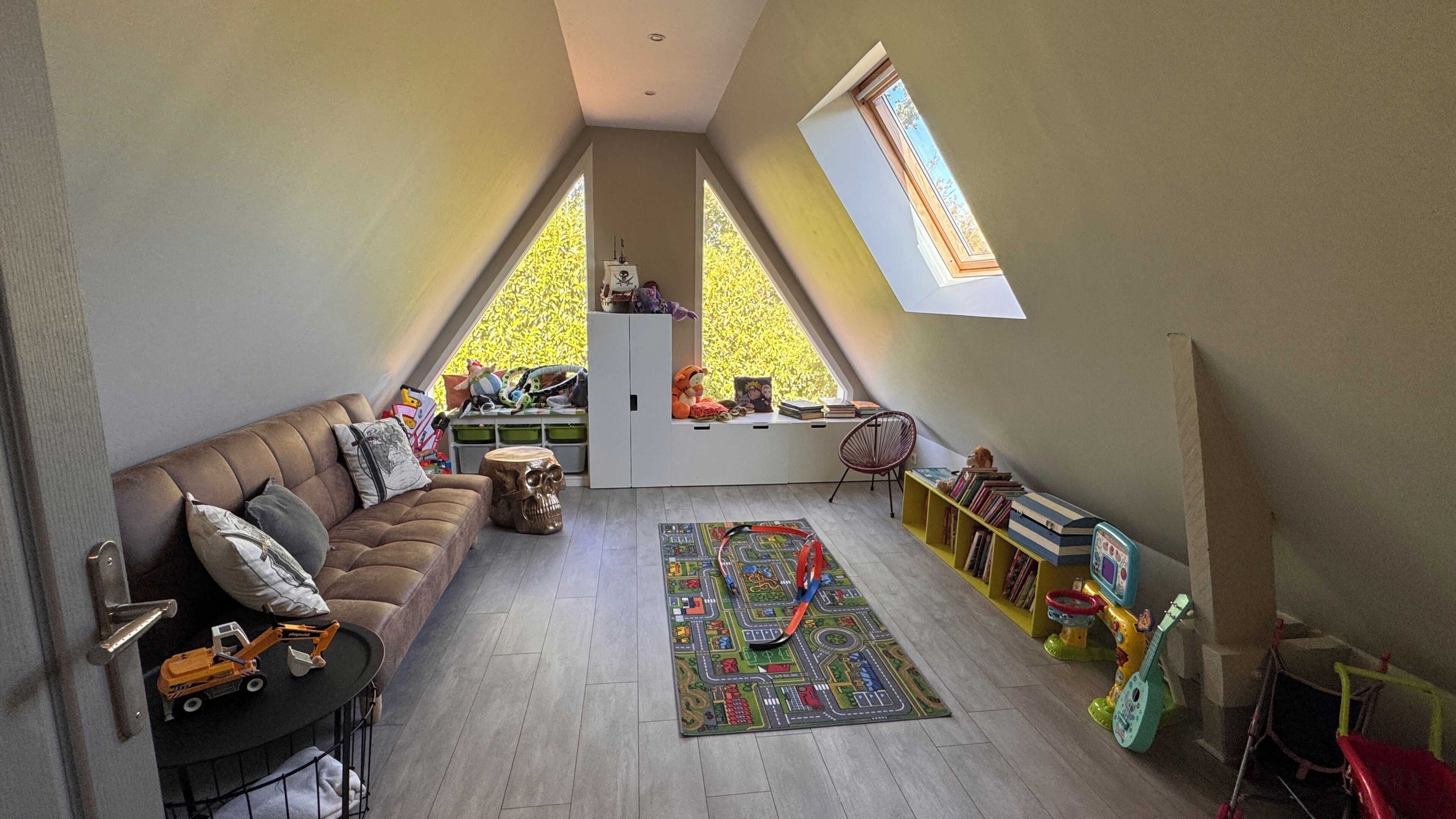 A loft-style room with a sloped ceiling, a brown leather sofa, a small table, a colorful rug, a bookshelf, and a window with a view of trees.