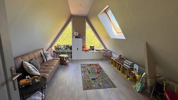 A loft-style room with a sloped ceiling, a brown leather sofa, a small table, a colorful rug, a bookshelf, and a window with a view of trees.
