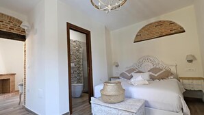 Un dormitorio con cama blanca, mesita de noche de madera, mesita auxiliar blanca y un arco de ladrillo.