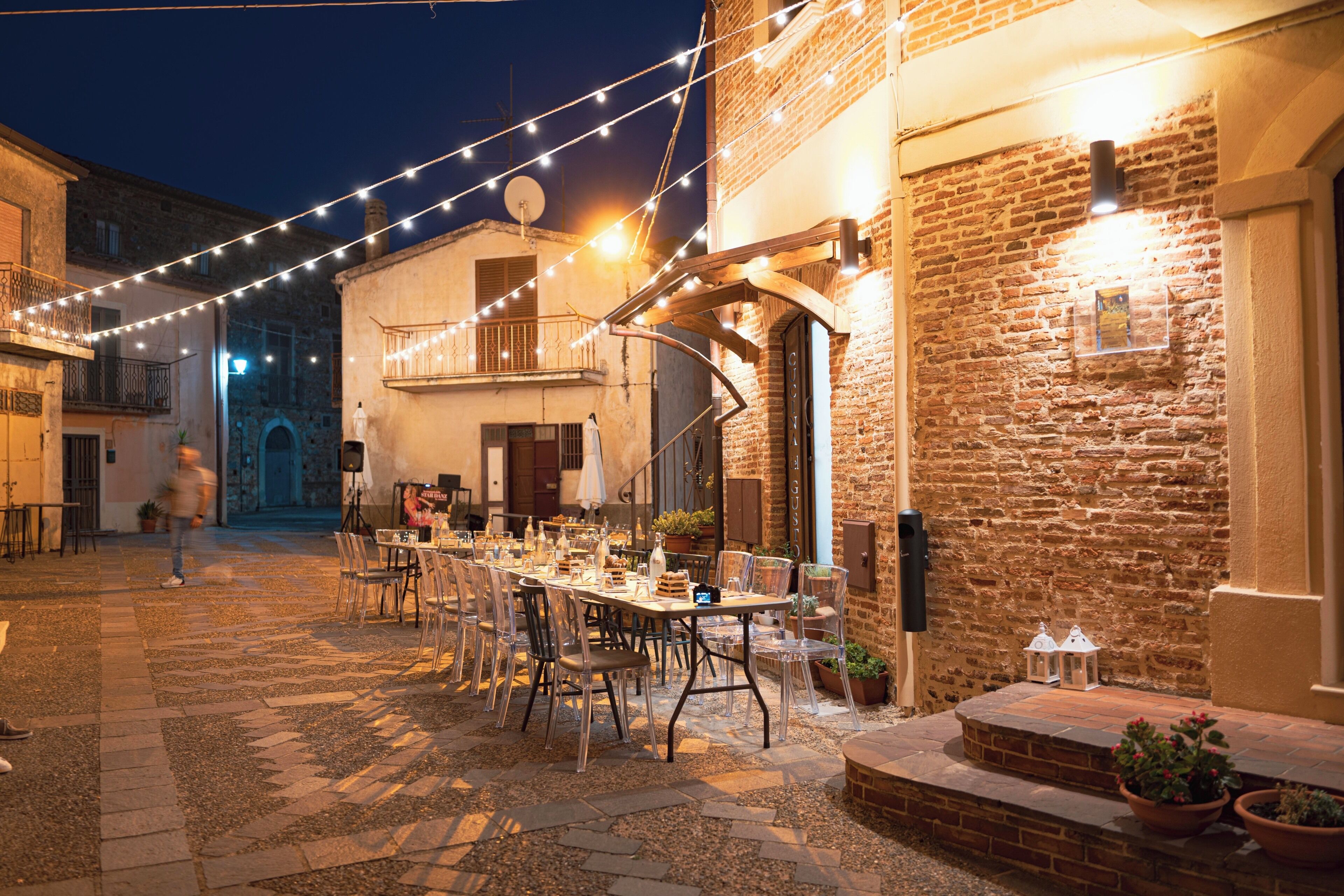 An outdoor dining area with tables and chairs, string lights, and a brick building.