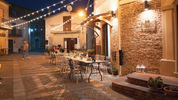 Un área de comedor al aire libre con mesas, sillas, luces colgantes y un edificio de ladrillo.