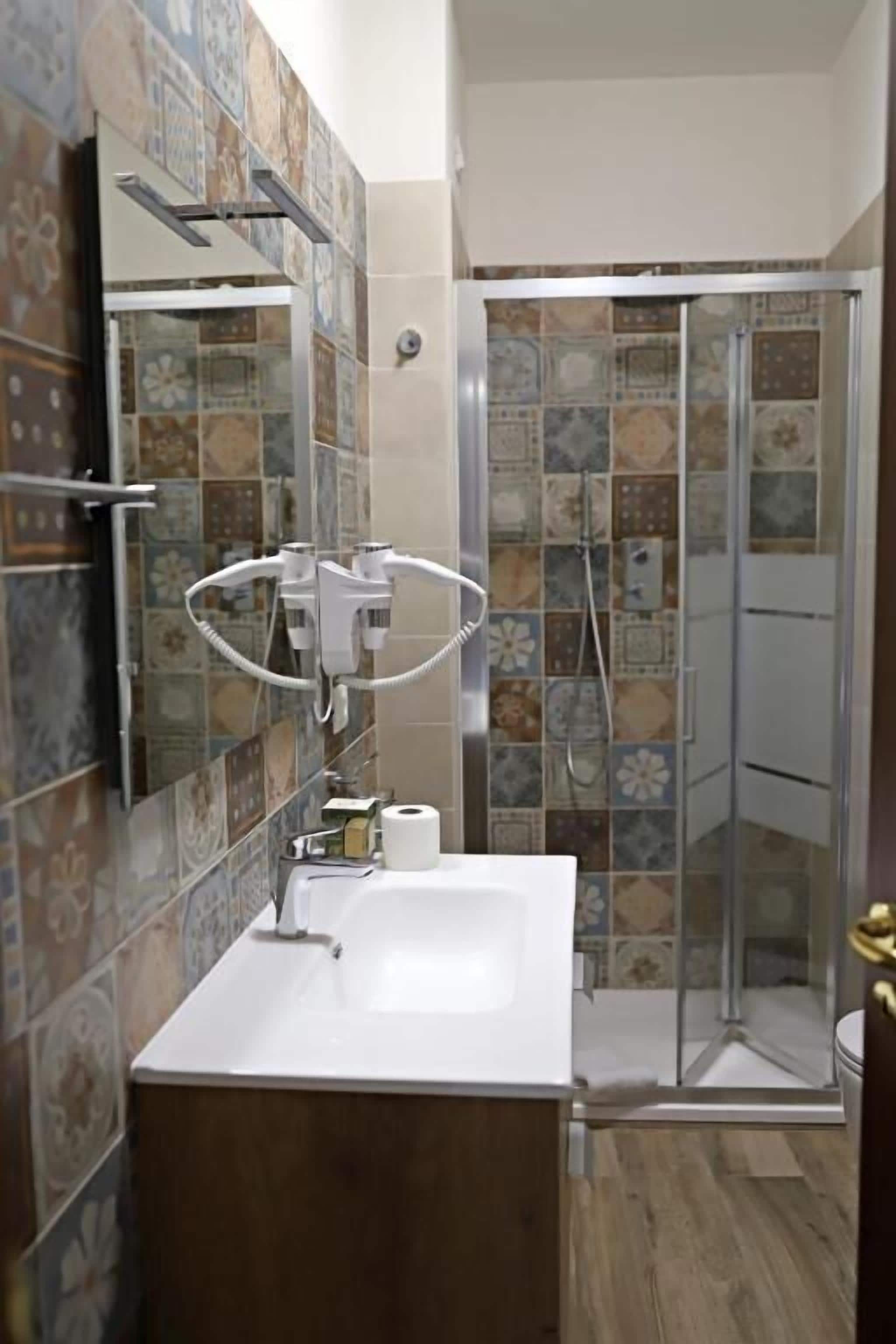 Ein Badezimmer mit einem weißen Waschbecken, einem Spiegel, einer Dusche mit Glastür und gemusterten Fliesen an den Wänden.