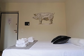 A hotel room with a bed, a wall-mounted pig illustration, a door, and a stack of towels on a table.