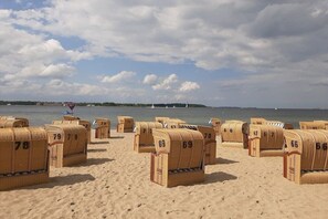 Des chaises de plage, numérotées de 67 à 79, sont disposées sur une plage de sable, offrant une vue sur la mer et un phare situé au loin.
