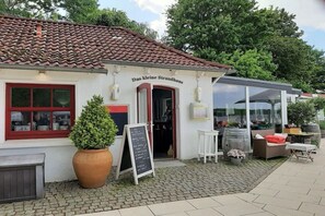Un petit bâtiment avec un toit en tuiles, une enseigne indiquant « Das kleine Strandhaus », et des sièges extérieurs avec des tables et des chaises.