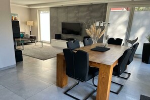 A modern dining area with a wooden table and black chairs, a flat-screen TV mounted on the wall, and a large window with blinds. - Seaside Lounge Rügen (Glowe)