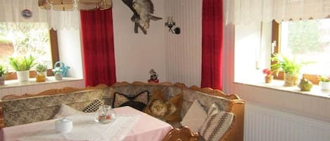Un comedor acogedor con un banco y mesa de madera, cortinas rojas y un adorno de pájaro colgado en la pared.