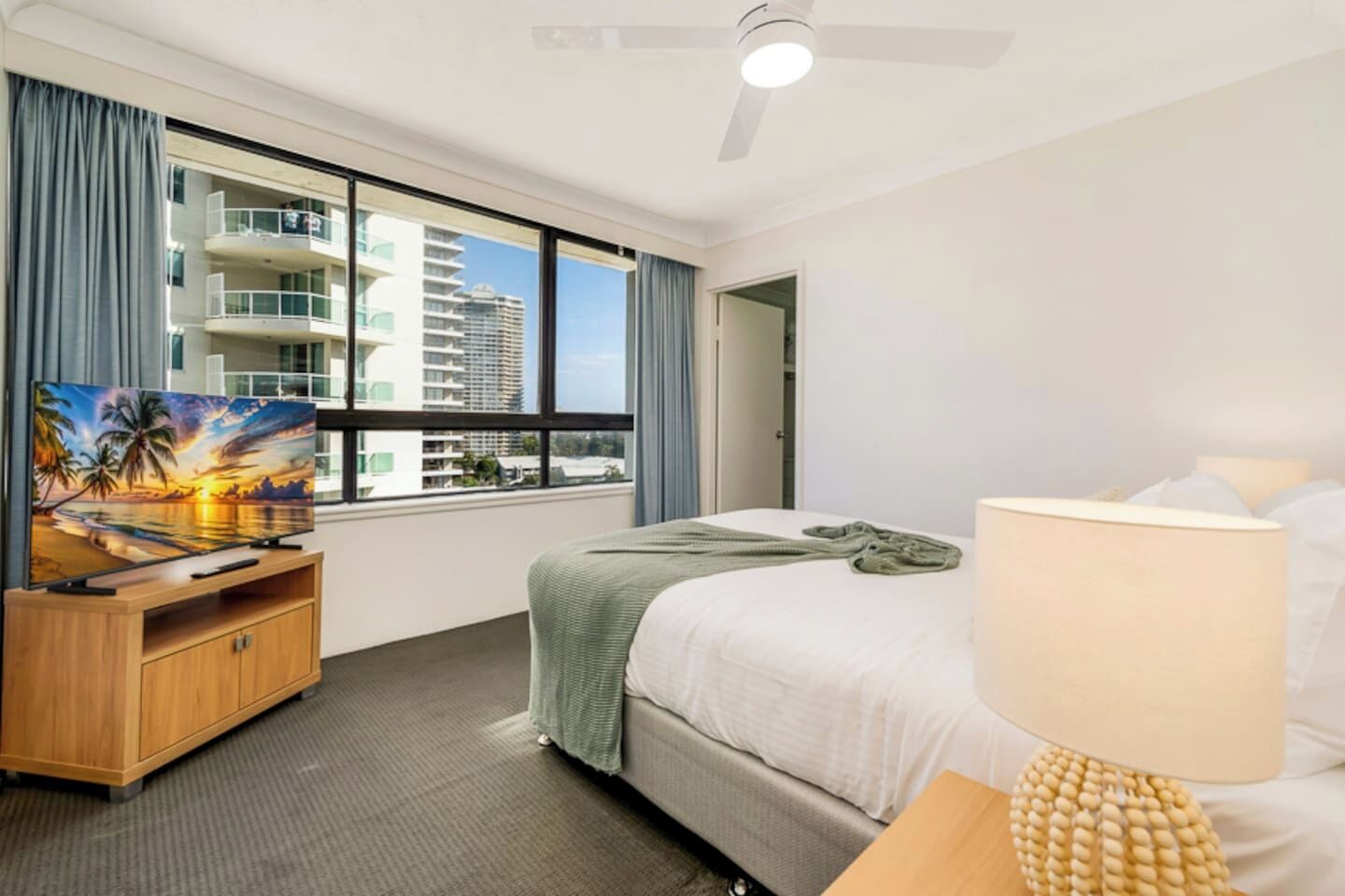 A hotel room with a bed, a TV on a wooden stand, a window with a view of buildings, a ceiling fan, and a lamp.