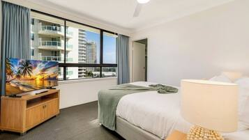 A hotel room with a bed, a TV on a wooden stand, a window with a view of buildings, a ceiling fan, and a lamp.