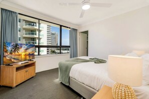 A hotel room with a bed, a TV on a wooden stand, a window with a view of buildings, a ceiling fan, and a lamp. - Carrington Getaway - Ocean Views, Parking, Pool (Main Beach)